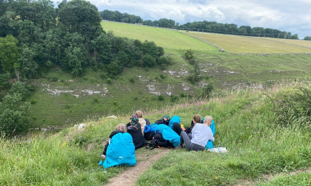 A group of students with camping gear taking a break on a path across a grassy hillside