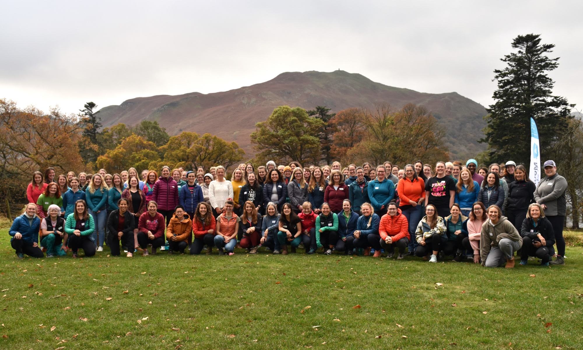 All attendees at the Women in Mountain Training Conference 2024 on the lawn outside Outward Bound Ullswater. Fell and autumn trees in the background.